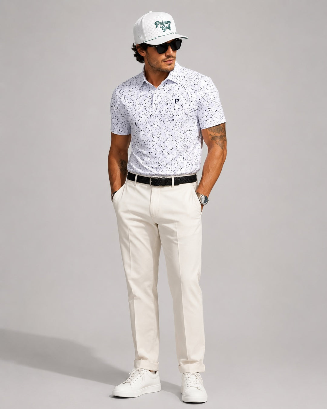 Man wearing a white patterned polo shirt, white pants, white sneakers, and a white cap with sunglasses on a gray background