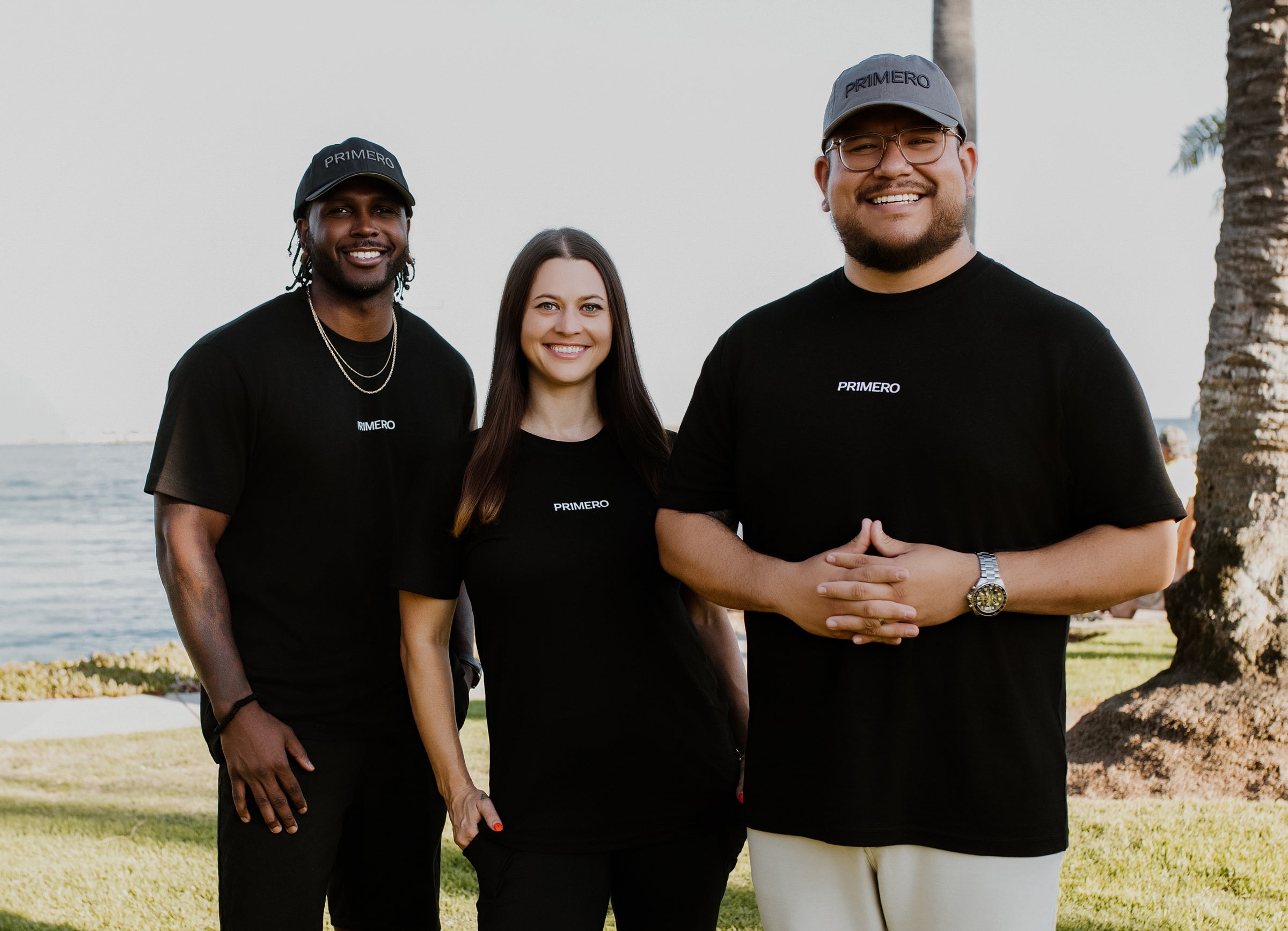 Three people wearing black shirts with a brand logo, standing outdoors near a palm tree and water.